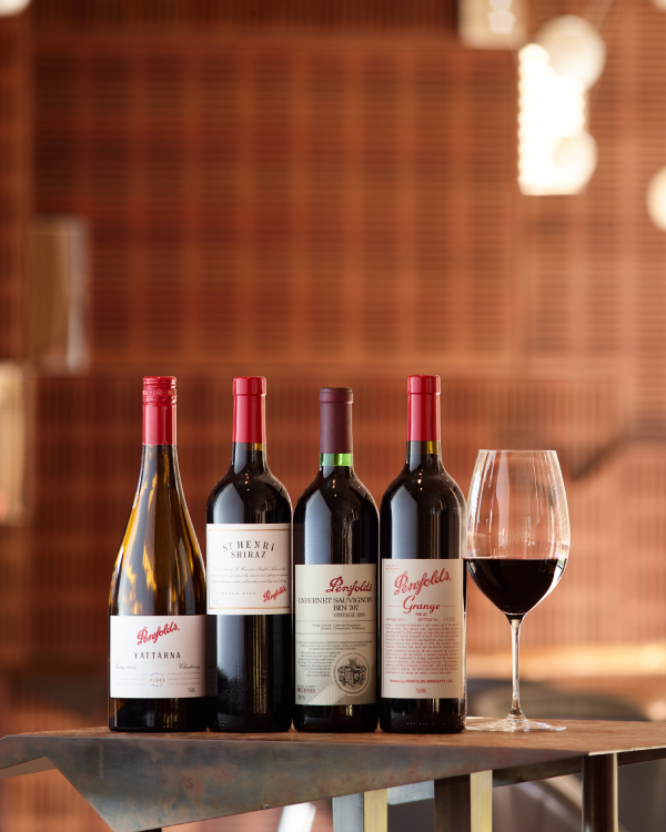 Four bottles of Penfolds wines stand on a cast iron table alongside a glass of red wine. 