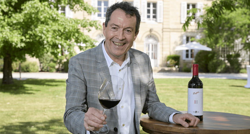 Peter Gago stands holding wine glass next to bottle of Penfolds II. Chateau visible behind