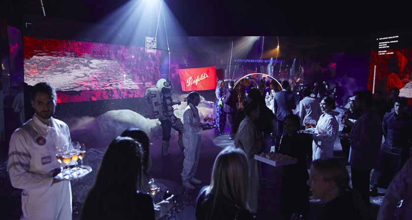 People mingle in a space themed room.  An astronaut stands with tray of champagne glasses