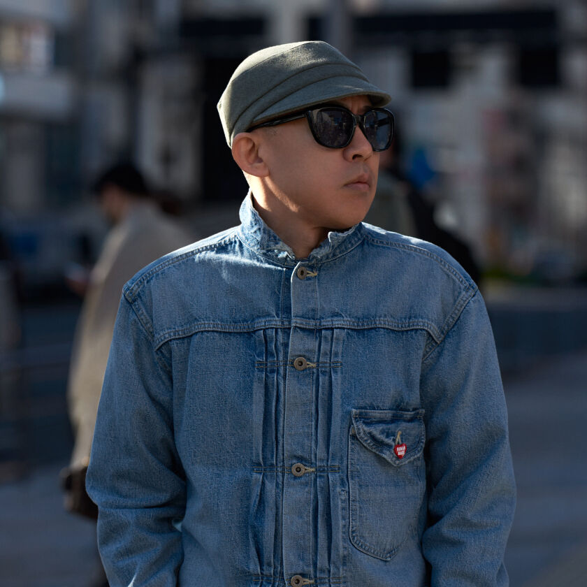 Nigo stands in blue denim jacket in the middle of a city. street